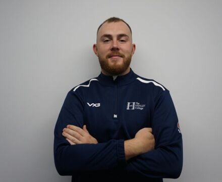A man with a beard stands confidently, arms crossed, wearing a navy sports jacket with a logo. The background is plain and light-colored.
